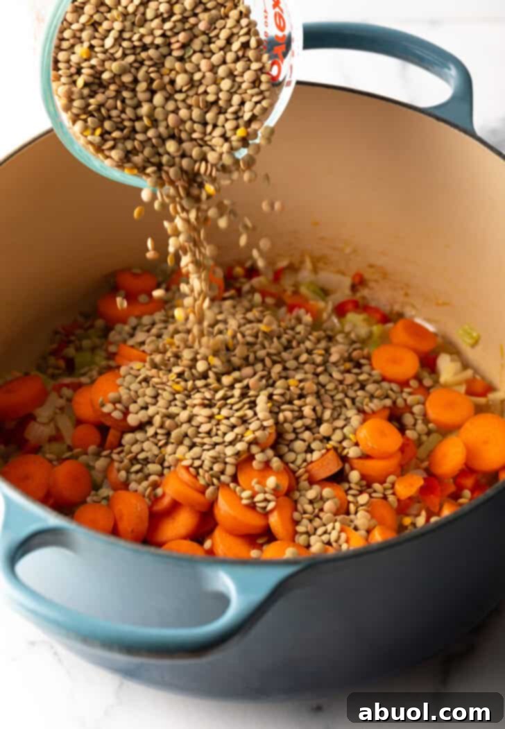Adding dried lentils from a clear measuring glass into a large cooking pot already containing sautéed carrots and other aromatic vegetables.