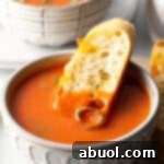 A white bowl of tomato soup with a slice of baguette in the soup, perfect for dipping.