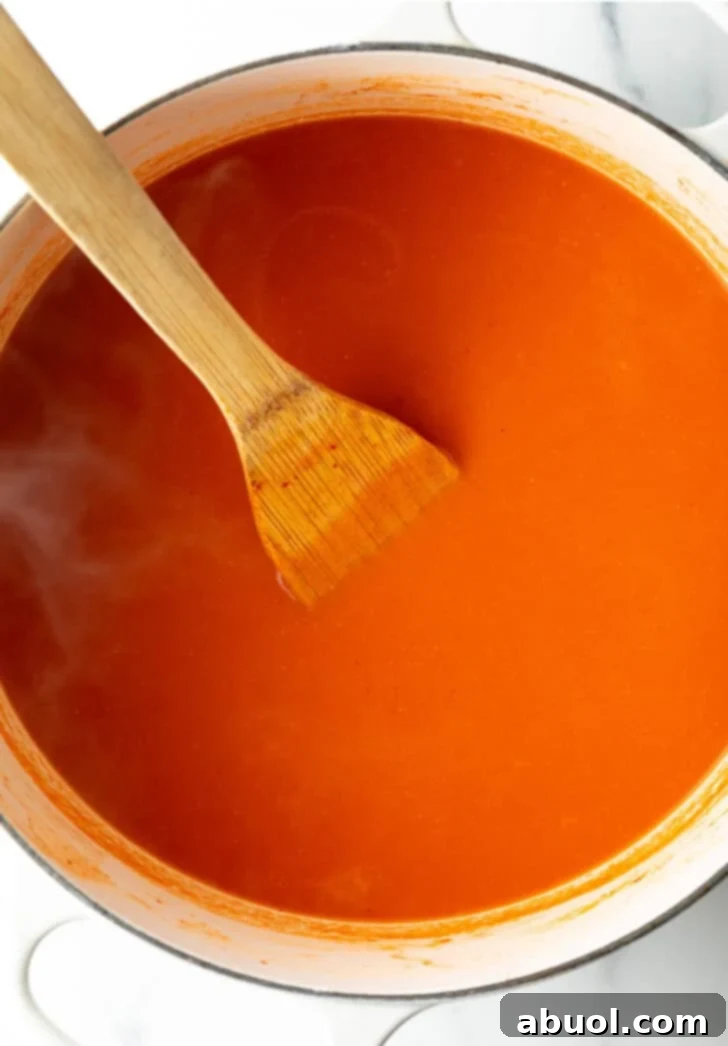 A top-down view captures a generous serving of vibrant, creamy tomato soup simmering gently in a large white pot, a wooden spatula resting within the rich, swirling liquid.