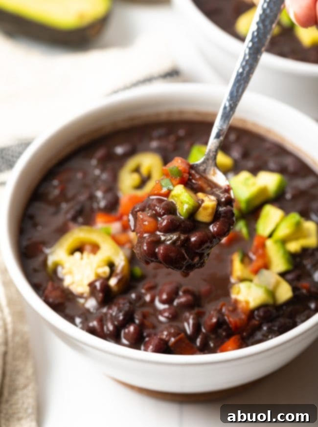 White bowl of black bean soup topped with slices of jalapenos, chopped avocado, and pico de gallo.