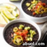 Two white bowls of black bean soup topped with slices of jalapenos, chopped avocado, and pico de gallo.