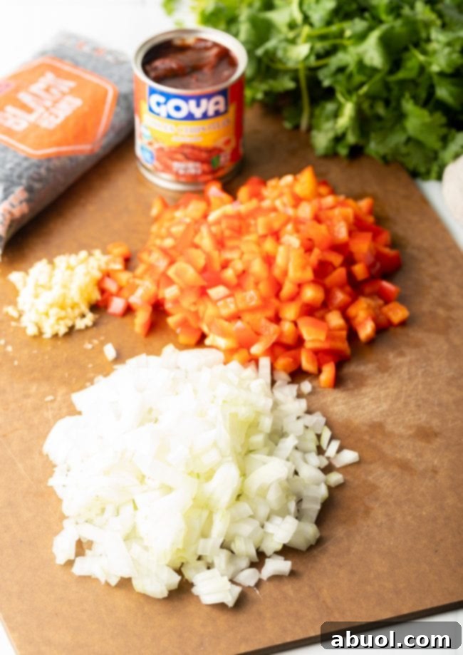 Cutting board with piles of chopped onion, chopped red peppers, garlic, and a can of ancho chile peppers, bag of black beans, and bunch of fresh cilantro.
