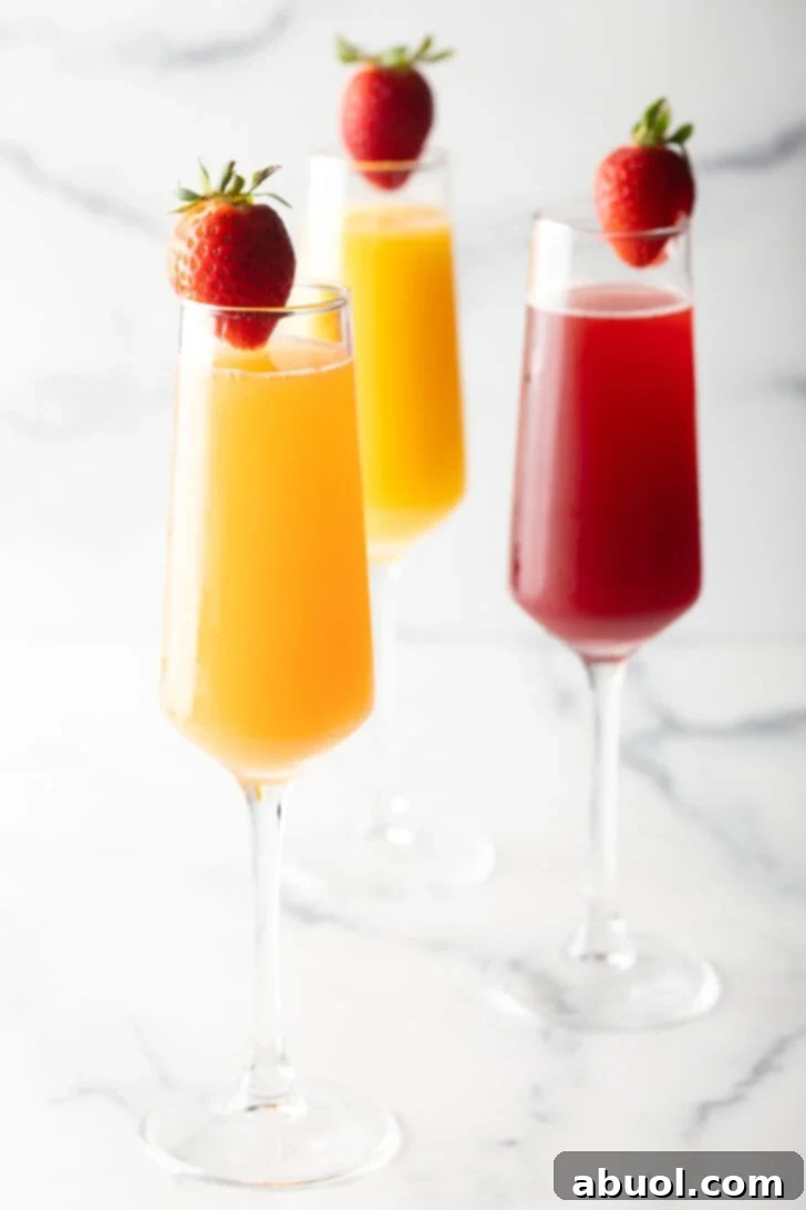 Two champagne flutes filled with orange mimosa, and another glass filled with a cranberry mimosa. Both glasses are garnished with a whole strawberry on the rim.