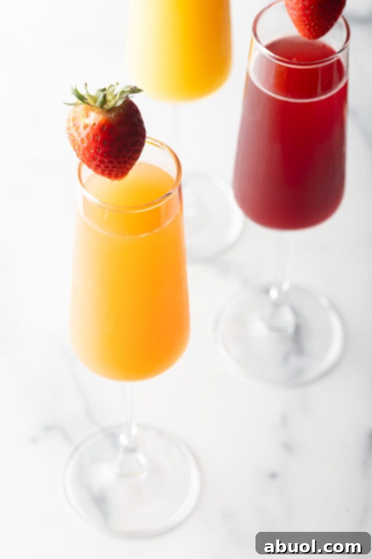 A champagne flute filled with orange mimosa, and another glass filled with a cranberry mimosa. Both glasses are garnished with a whole strawberry on the rim.