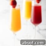 Two champagne flutes filled with orange mimosa, and another glass filled with a cranberry mimosa. Both glasses are garnished with a whole strawberry on the rim.