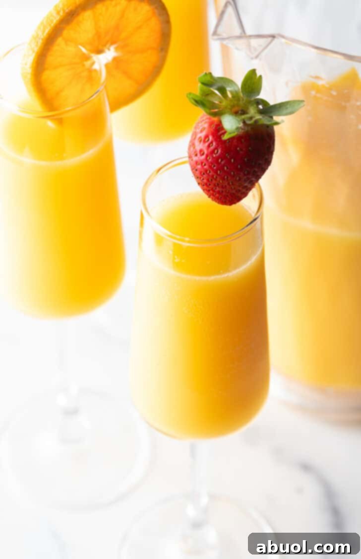 Two glasses of mimosas, one garnished with a large strawberry on the rim, the other garnished with an orange slice on the rim.
