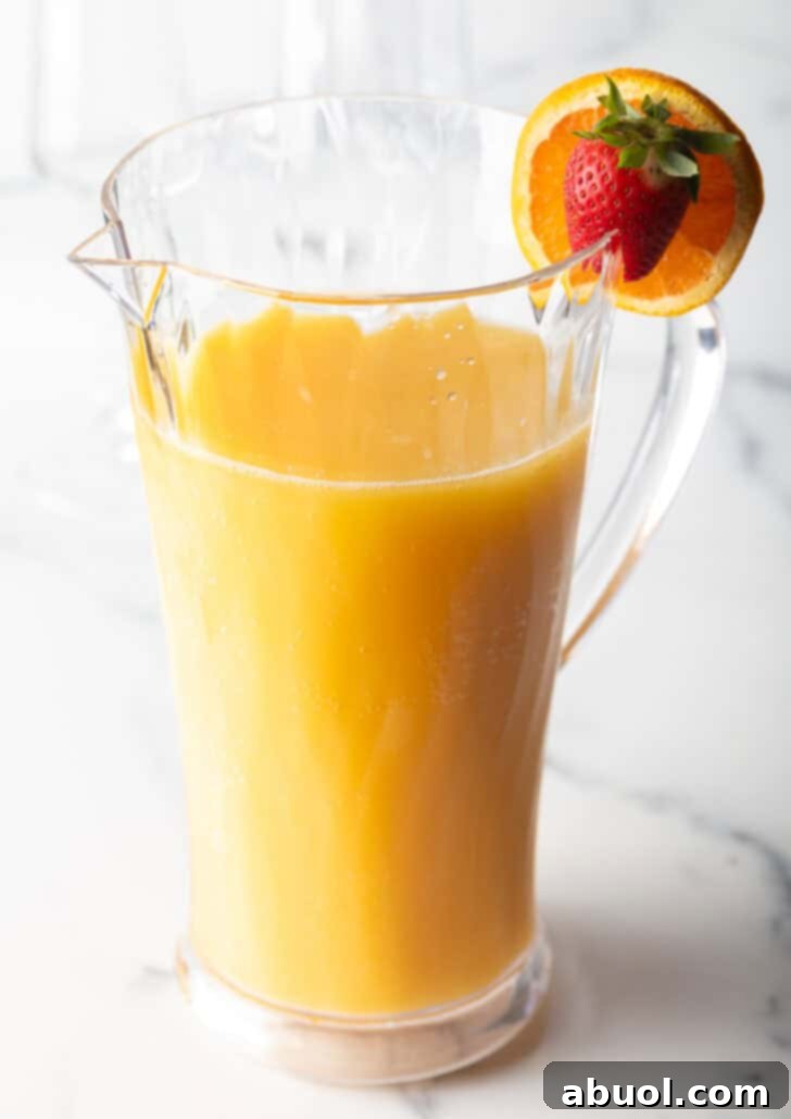 Large glass pitcher of mimosa with an orange and strawberry garnish on the rim.