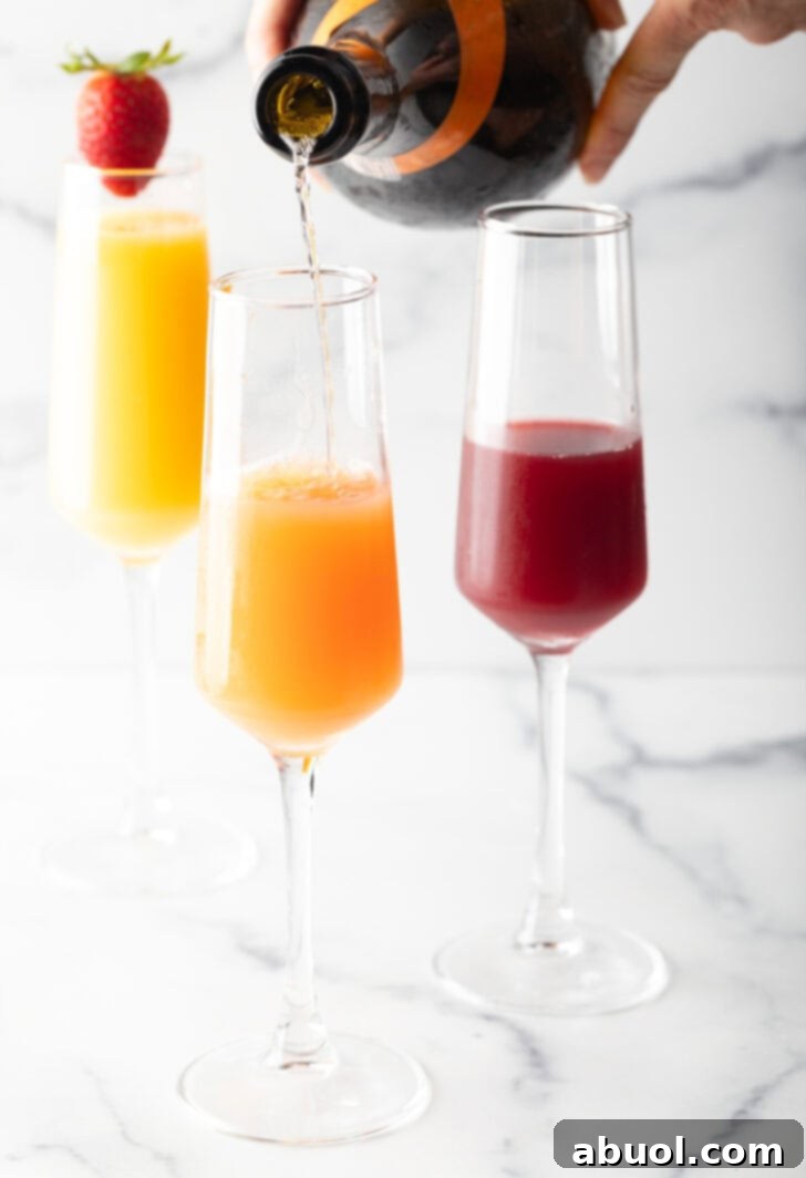 3 champagne glass against a white marble backdrop. The glass in the background is full of orange juice and garnished with a whole strawberry on the rim. In the foreground are a glass half full with cranberry juice, and a glass half full of orange juice with sparkling wine being poured in to the glass.