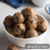 White bowl brimming with energy balls that are speckled with chocolate chips and raisins. One ball is on the table next to the bowl.