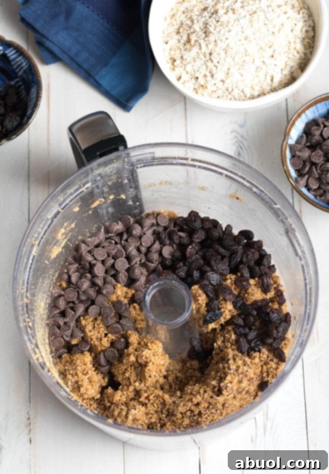 Vegan chocolate chips being gently added to the crumbled no-bake oatmeal cookie dough in a food processor, ready for a final mix by hand.