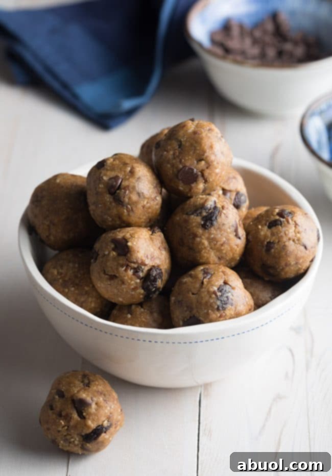 A white bowl brimming with perfectly rounded energy balls, generously speckled with rich chocolate chips and sweet raisins. One energy ball rests enticingly on the table beside the bowl.