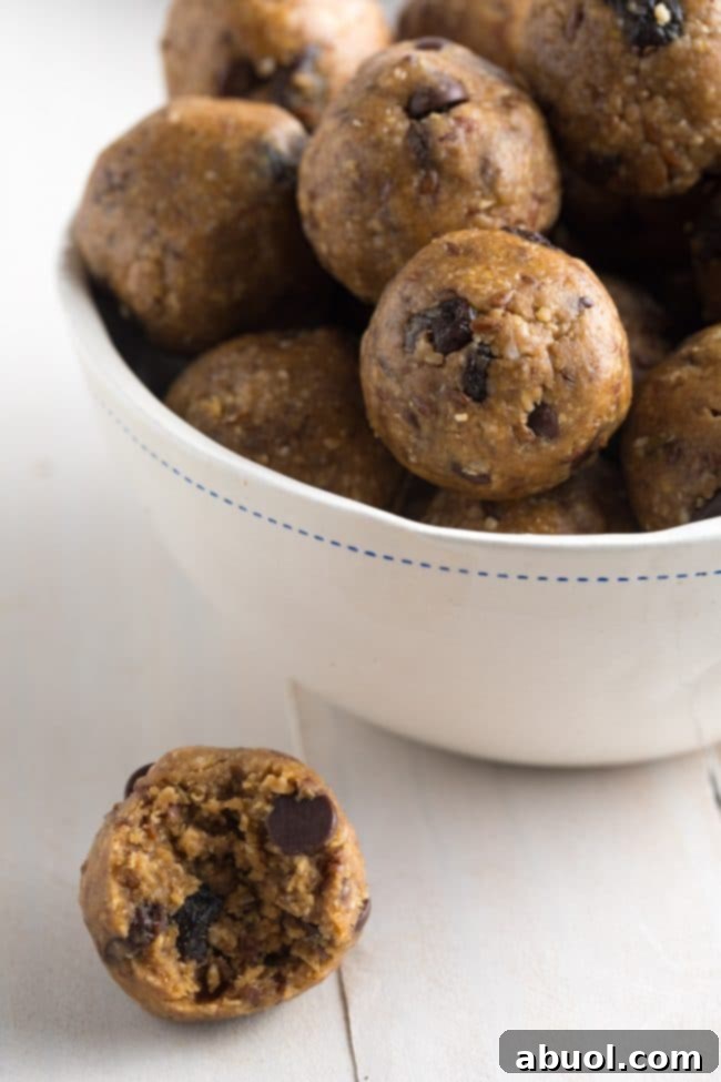 A white bowl brimming with energy balls speckled with chocolate chips and raisins. One energy ball with a bite taken out of it rests on the table, showcasing its delicious texture.