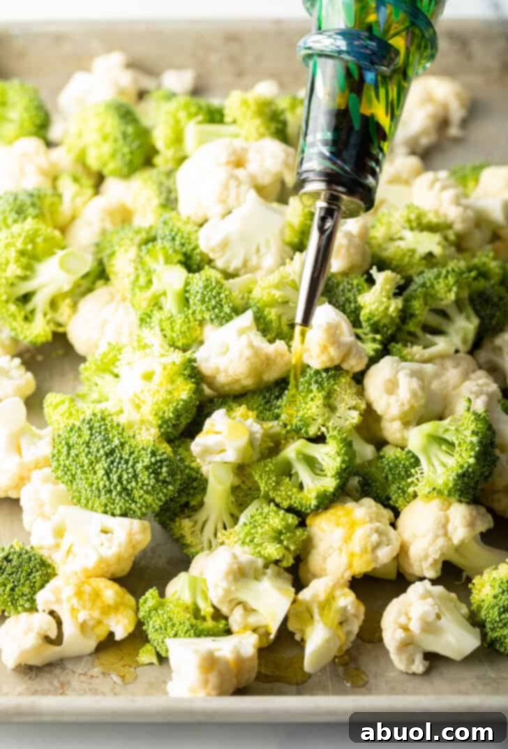 A close-up shot of raw broccoli and cauliflower florets evenly spread on a baking sheet, being generously drizzled with olive oil.