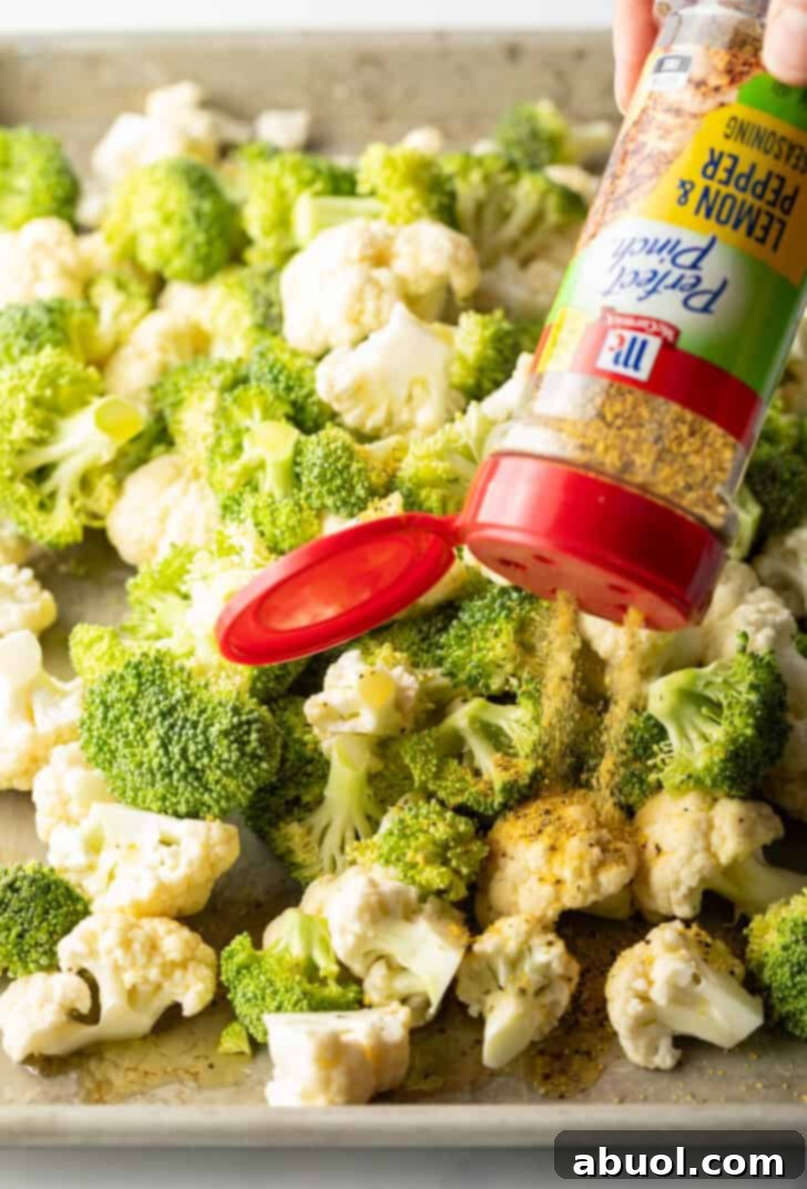Lemon pepper seasoning being sprinkled liberally over the olive-oil-coated broccoli and cauliflower florets on a baking sheet, preparing them for roasting.
