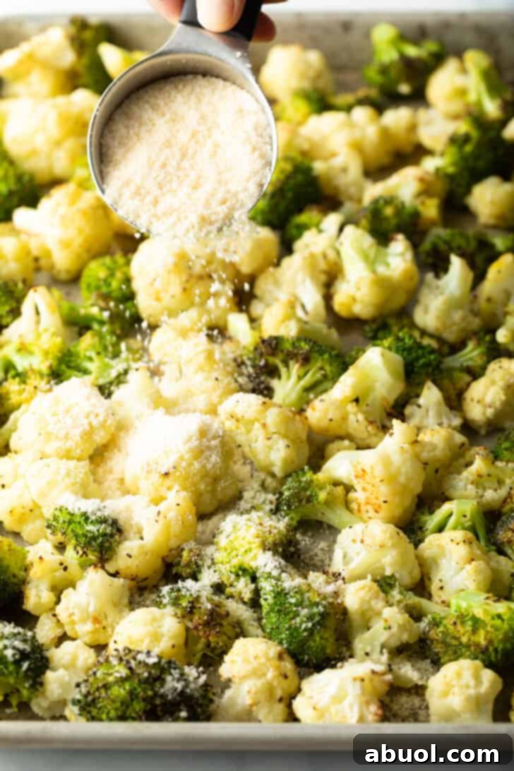Grated Parmesan cheese being evenly sprinkled over a baking sheet filled with partially roasted broccoli and cauliflower, adding the final touch before returning to the oven.
