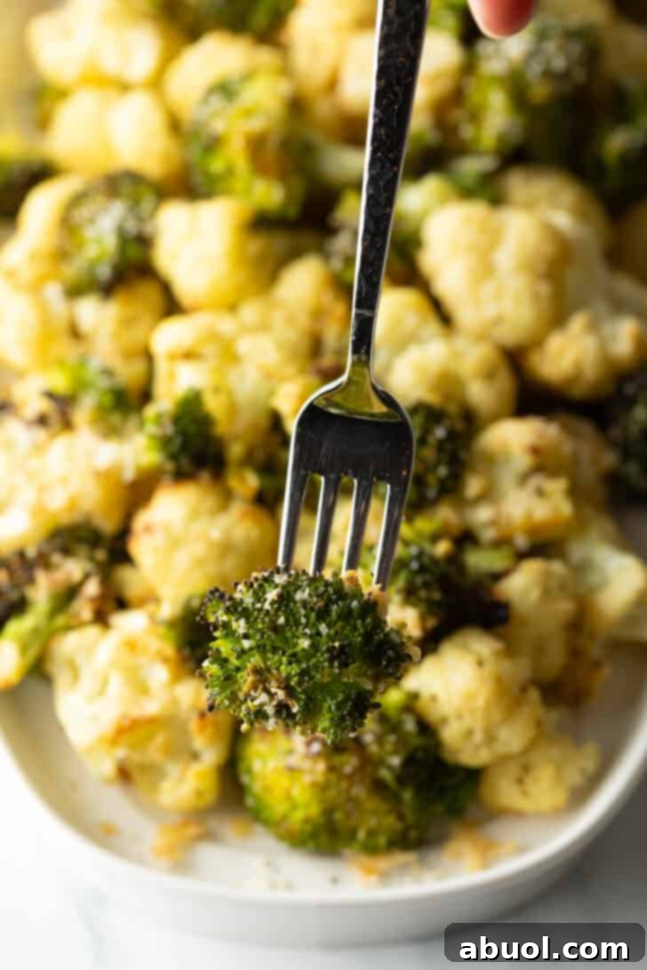 A fork proudly holds a perfectly roasted piece of broccoli towards the camera, with a blurred background of a platter overflowing with more roasted cauliflower and broccoli florets.