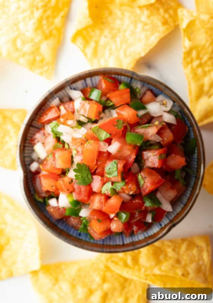 Top down view of a white bowl of fresh pico de gallo with chopped tomatoes, onion, jalapeno, and cilantro. The bowl is surrounded by tortilla chips, ready for dipping.