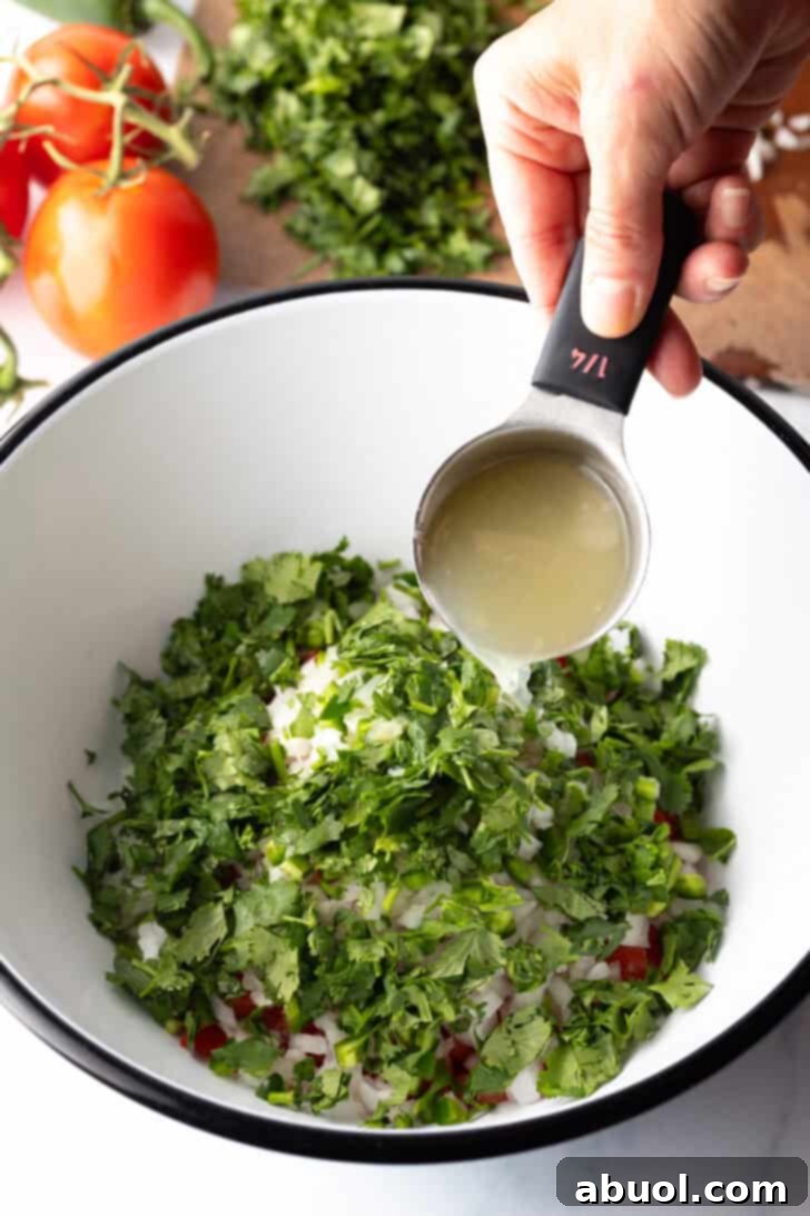 Fresh chopped cilantro has been added to the bowl. A hand is pouring lime juice from a small metal measuring cup into the mixture.