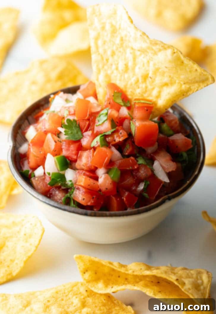 A white bowl of fresh pico de gallo with chopped tomatoes, onion, jalapeno, and cilantro. A tortilla chip is sticking out of the bowl, ready to be enjoyed.