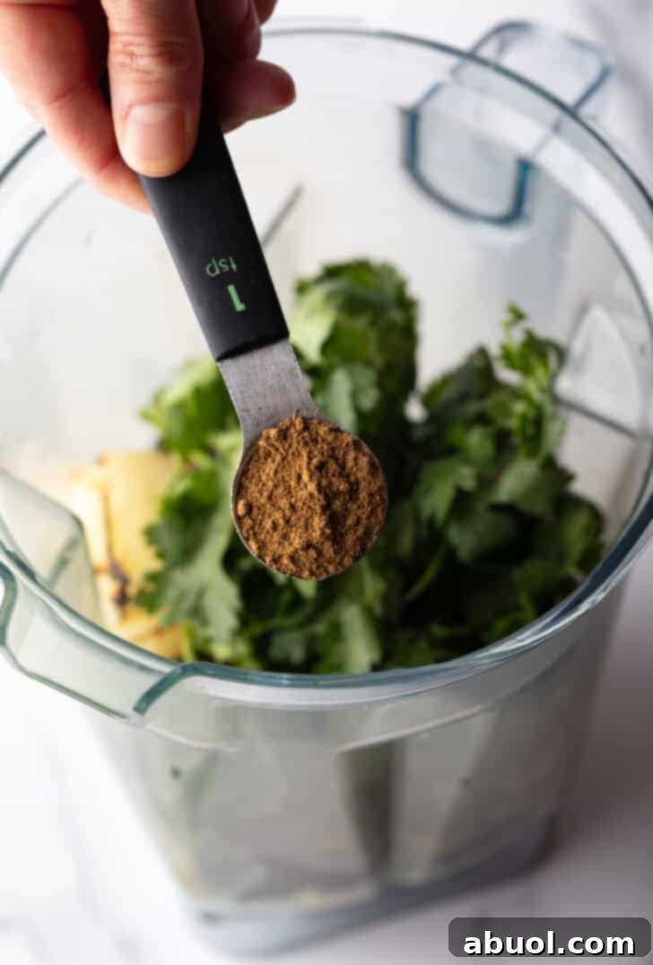 A hand pouring ground cumin into a blender pitcher filled with vibrant green herbs and roasted vegetables.