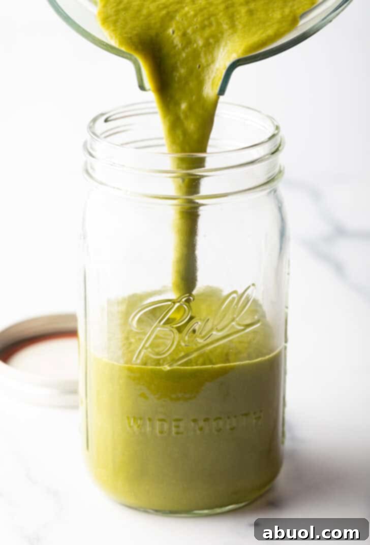 Pouring the freshly blended green sauce from a clear pitcher into a large, wide-mouth glass mason jar for storage.
