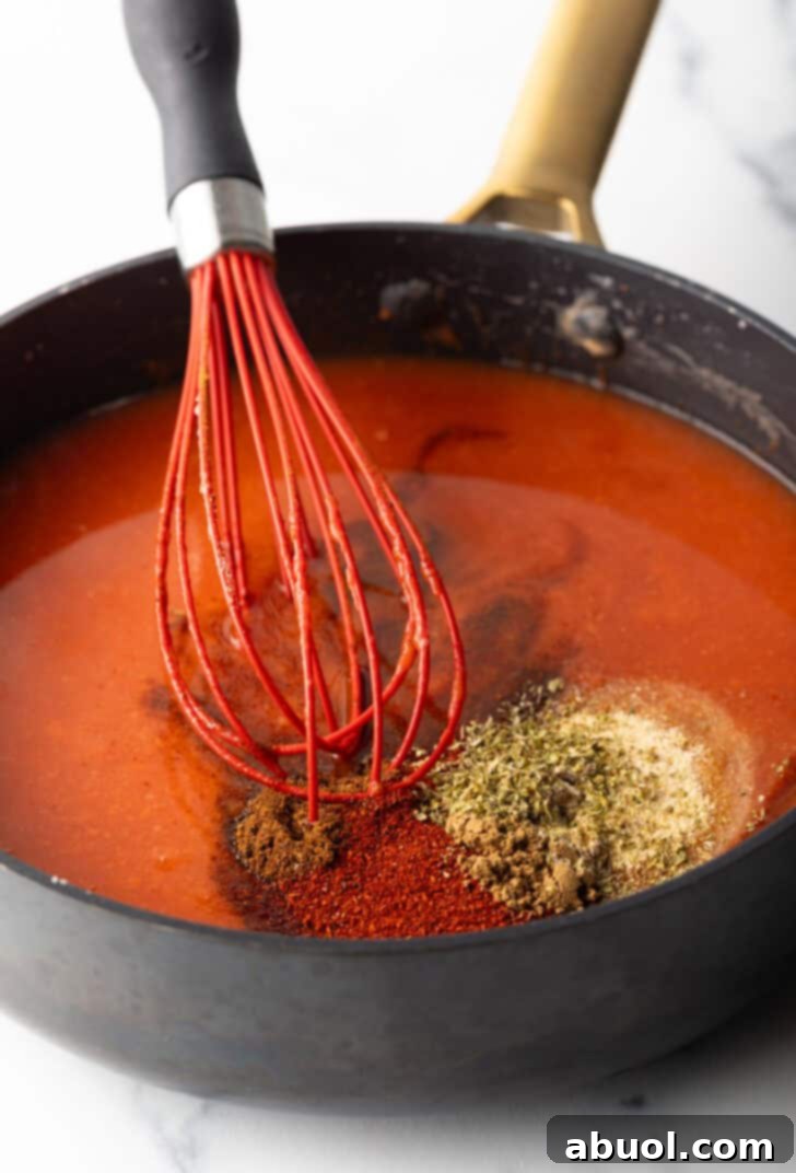 Large deep skillet with red sauce, spices, and a red whisk.