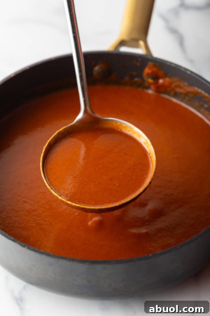 Metal ladleful of red sauce being held above a black pan of sauce.