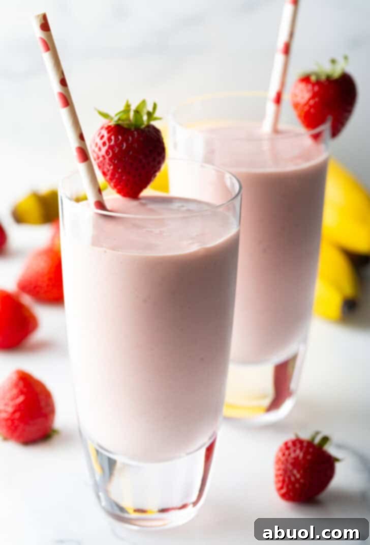 Two tall glasses of strawberry banana smoothies, each garnished with a whole strawberry on the rim and a red and white straw.