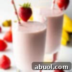 Two tall glasses of strawberry banana smoothies, each garnished with a whole strawberry on the rim and a red and white straw.