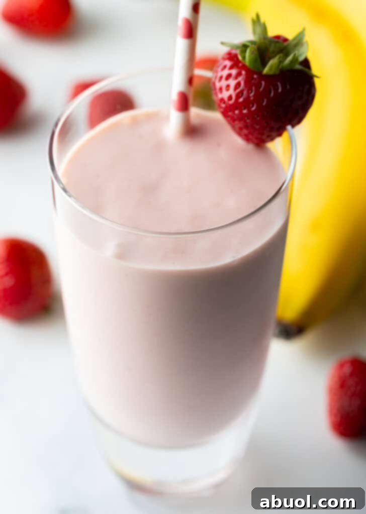 Tall glass of strawberry and banana smoothie, garnished with a whole strawberry on the rim and a red and white straw.