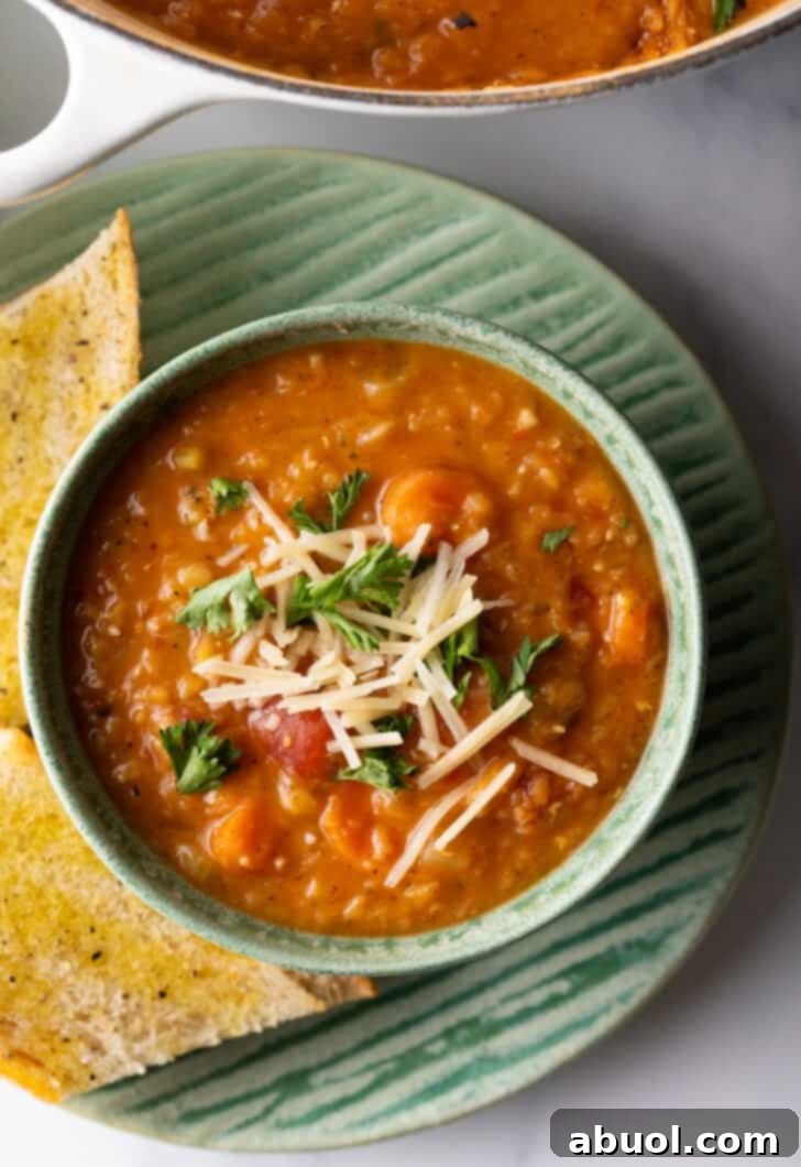 An inviting top-down view of a green bowl brimming with rich red lentil soup. The soup is beautifully garnished with fresh chopped herbs and a hint of shredded cheese. The bowl is set on a simple plate, accompanied by two golden-brown baguette slices, creating a scene of comforting deliciousness.