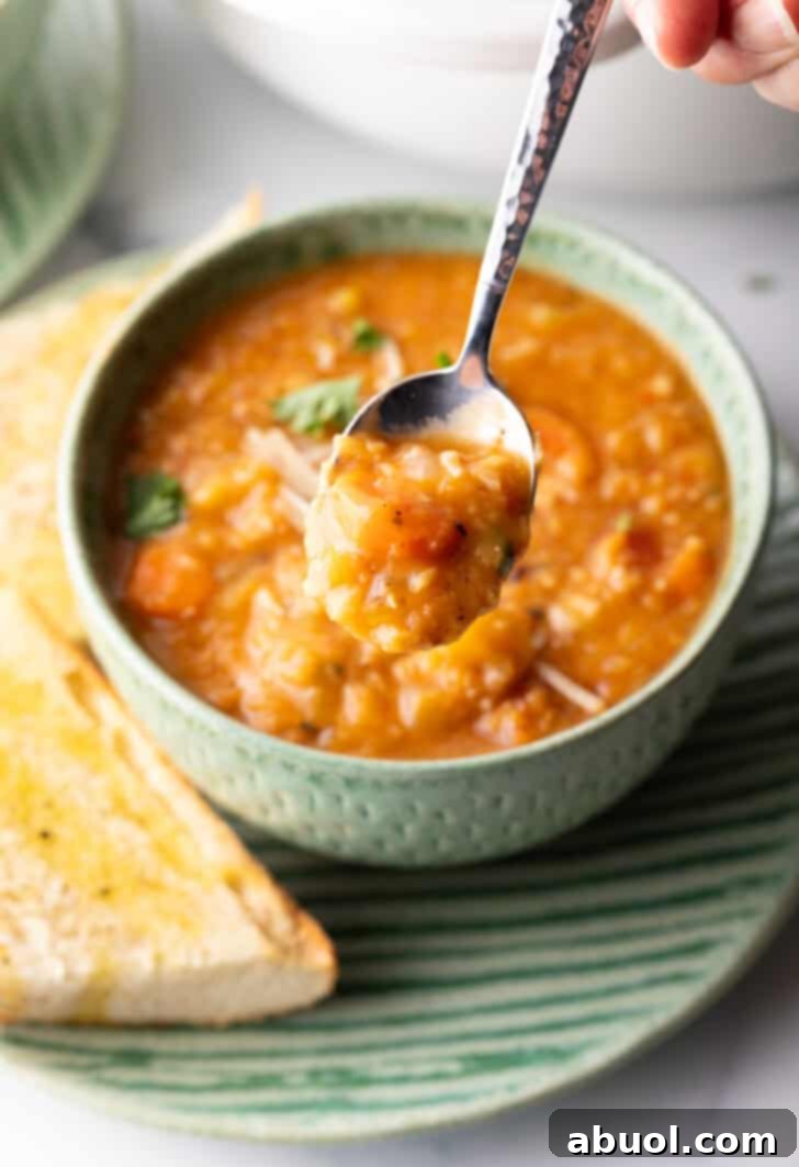 A hand gracefully holds a spoonful of rich, red lentil soup, bringing it closer to the camera to showcase its inviting texture and warmth. The background remains softly blurred, keeping the focus on the delicious, rustic soup.