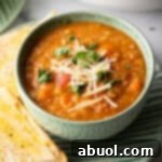 A green bowl of red lentil soup topped with chopped herbs and shredded cheese. The bowl is on a plate with two baguette slices.