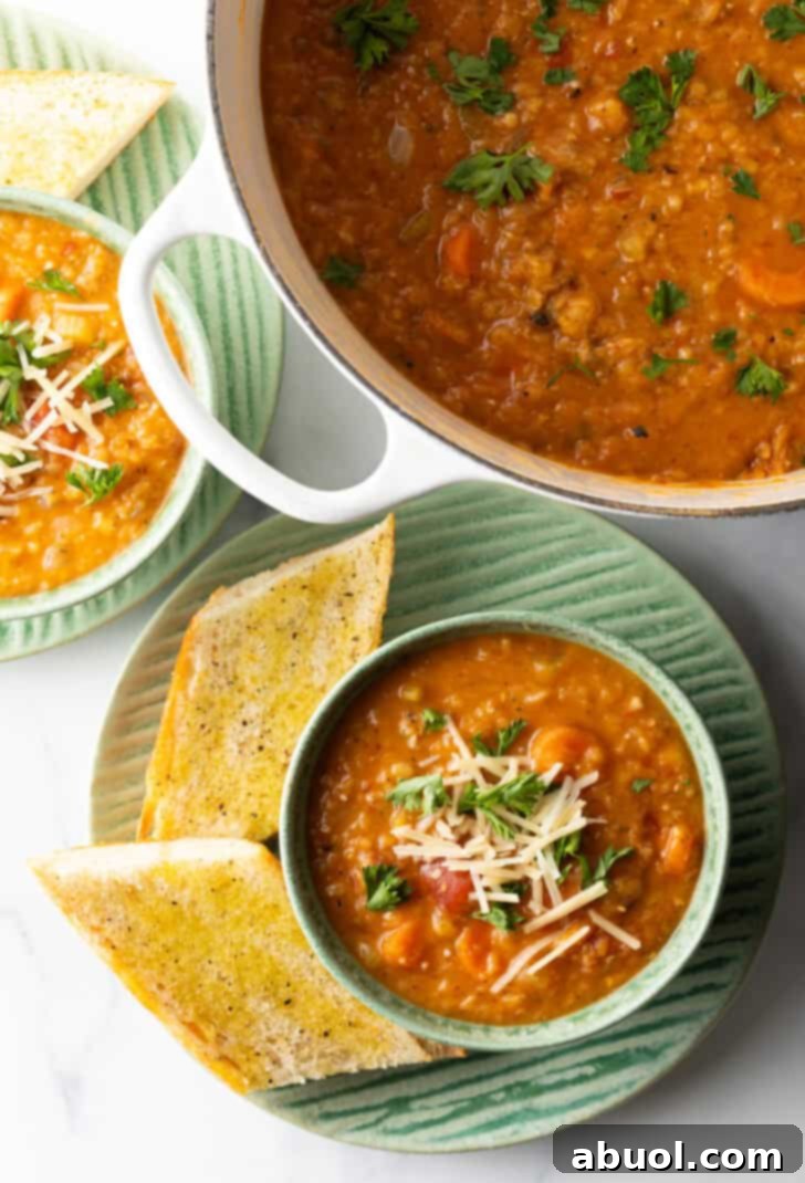 An overhead view of a vibrant green bowl filled with warm carrot and red lentil soup. The surface is adorned with a generous sprinkle of fresh chopped herbs and a touch of shredded cheese, creating an inviting contrast. The bowl is presented on a simple plate, flanked by two golden, toasted slices of baguette, emphasizing its rustic and comforting appeal.