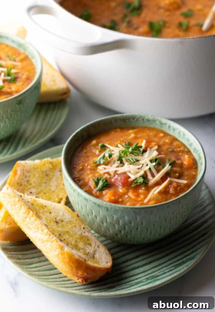 A beautifully presented green bowl of comforting carrot red lentil soup. The soup is generously topped with vibrant chopped herbs and a delicate sprinkle of shredded cheese. The bowl rests on a rustic plate, perfectly complemented by two slices of toasted baguette, creating an inviting and wholesome meal scene.