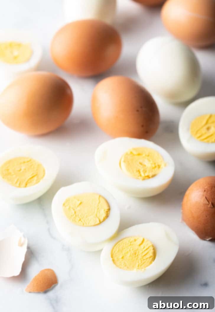 A dozen perfectly hard boiled eggs on a pristine white marble background, with some peeled halves showing vibrant yellow yolks and some brown eggs still in their shells, alongside one fully peeled white egg.