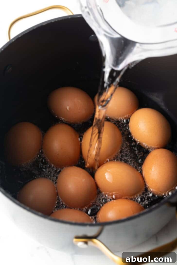 Water being carefully poured from a clear glass measuring cup over a dozen brown eggs in a black cooking pot, ensuring full submersion.