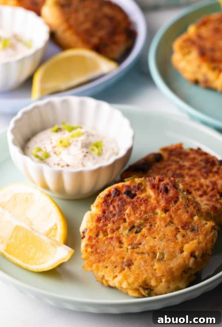 Two golden-brown, crispy salmon patties resting on a vibrant green plate, accompanied by a small white ramekin filled with creamy tartar sauce and two bright lemon wedges, ready to be served.