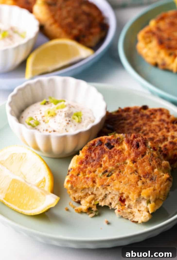 A single salmon patty on a green plate, with a bite taken out of it, showcasing its moist, flaky interior and crispy golden crust, alongside tartar sauce and lemon wedges.