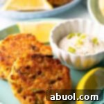 Two salmon patties on a green plate, with a white ramekin of tartar sauce and two lemon wedges.