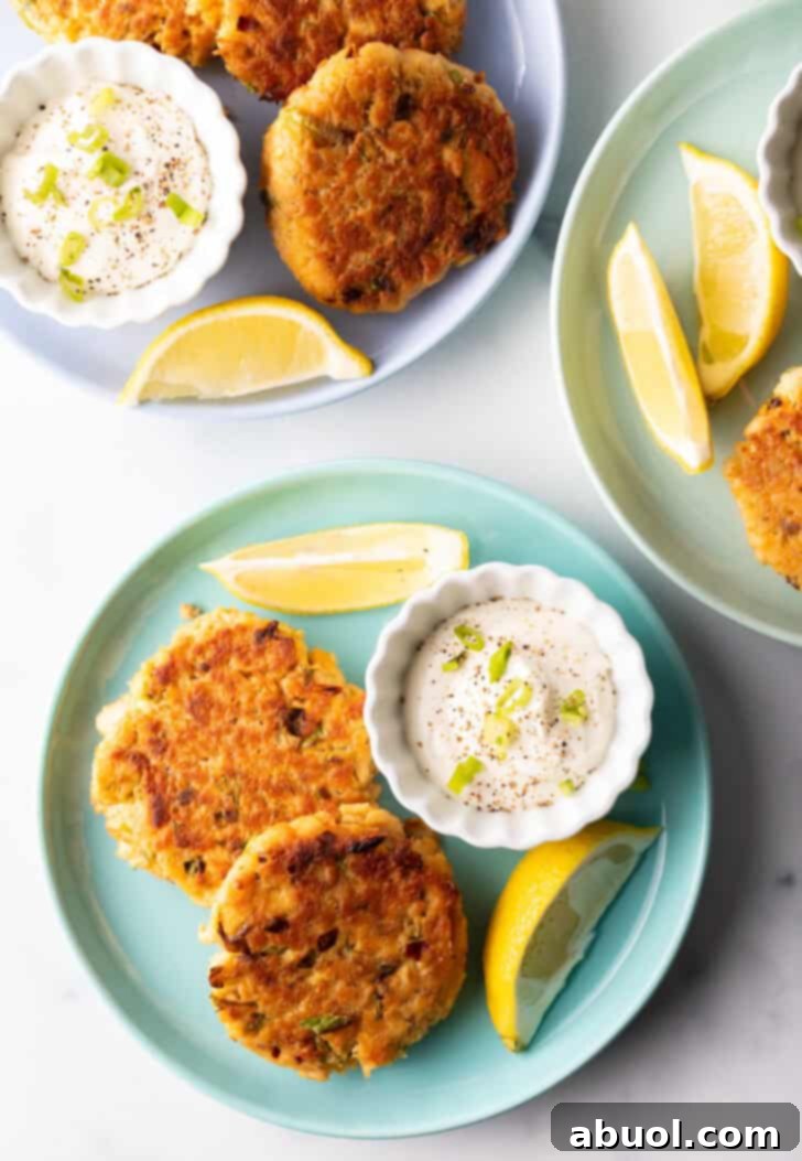 Two perfectly pan-fried salmon patties on a green plate, with a side of creamy tartar sauce in a white ramekin and two vibrant lemon wedges, emphasizing their golden-brown crust and inviting presentation.