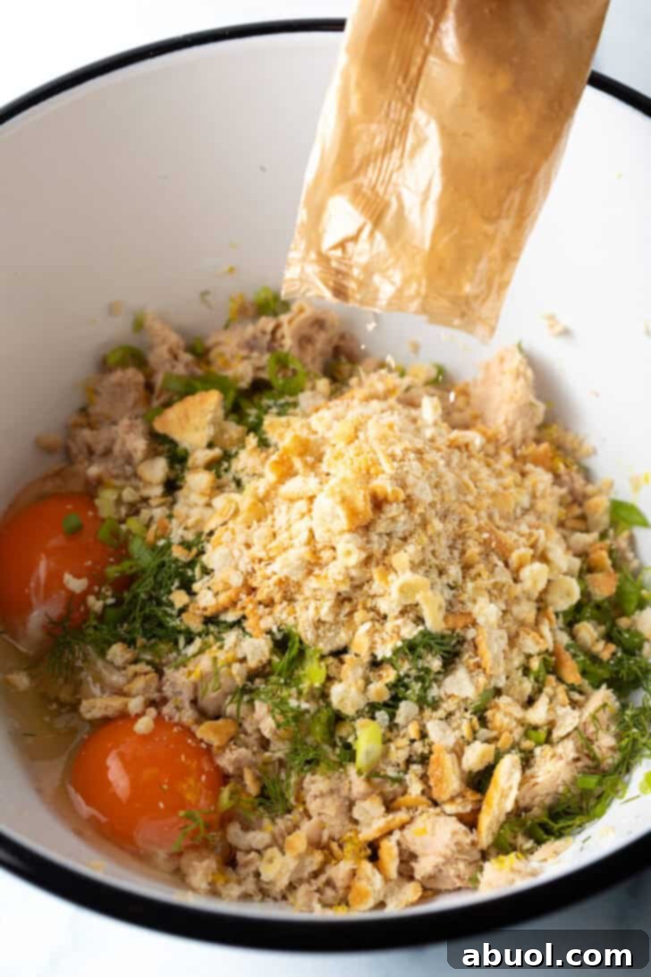 A large white mixing bowl showcases a vibrant mixture of flaked canned salmon, coarse breadcrumbs, finely chopped fresh herbs, and cracked eggs, ready to be combined.