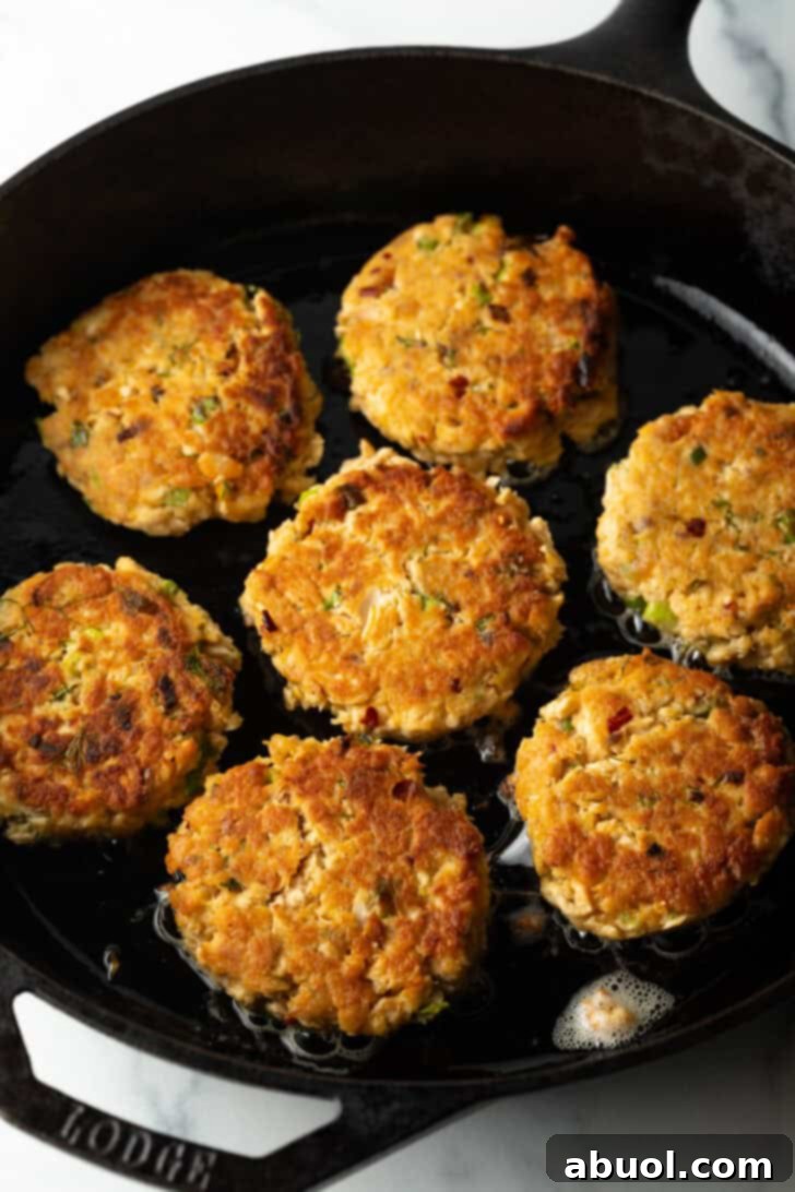 A top-down view captures seven golden-brown salmon patties sizzling and cooking to perfection in a well-seasoned cast iron skillet.