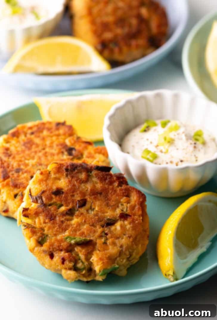 A close-up shot of two salmon patties on a green plate, perfectly cooked with a golden crust, served with creamy tartar sauce and fresh lemon wedges, highlighting their appetizing texture.