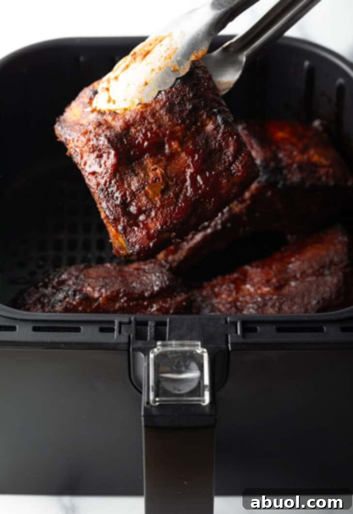 Tongs carefully lifting a perfectly cooked, sauce-glazed rib from an air fryer basket, with another juicy rib visible in the background.