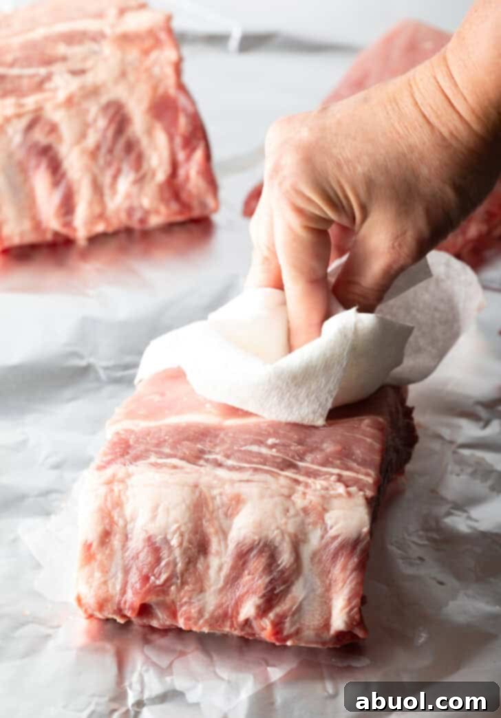 A hand using a paper towel to pat dry a small rack of baby back ribs, essential for proper seasoning adherence.