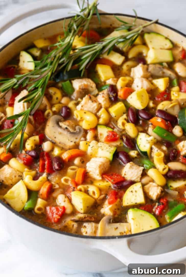 A large white pot filled to the brim with vibrant chicken minestrone soup, garnished with fresh sprigs of rosemary, ready to serve.