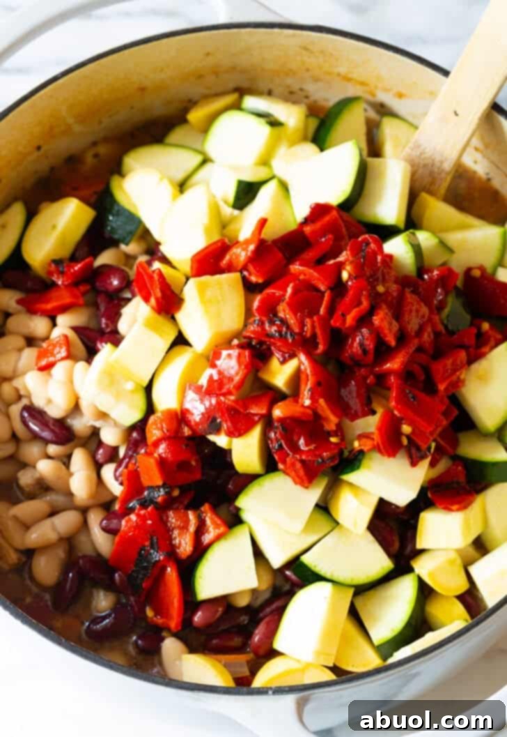 Hand adding freshly chopped roasted red peppers to a simmering pot filled with a colorful mix of chopped zucchini and carrots for minestrone soup.
