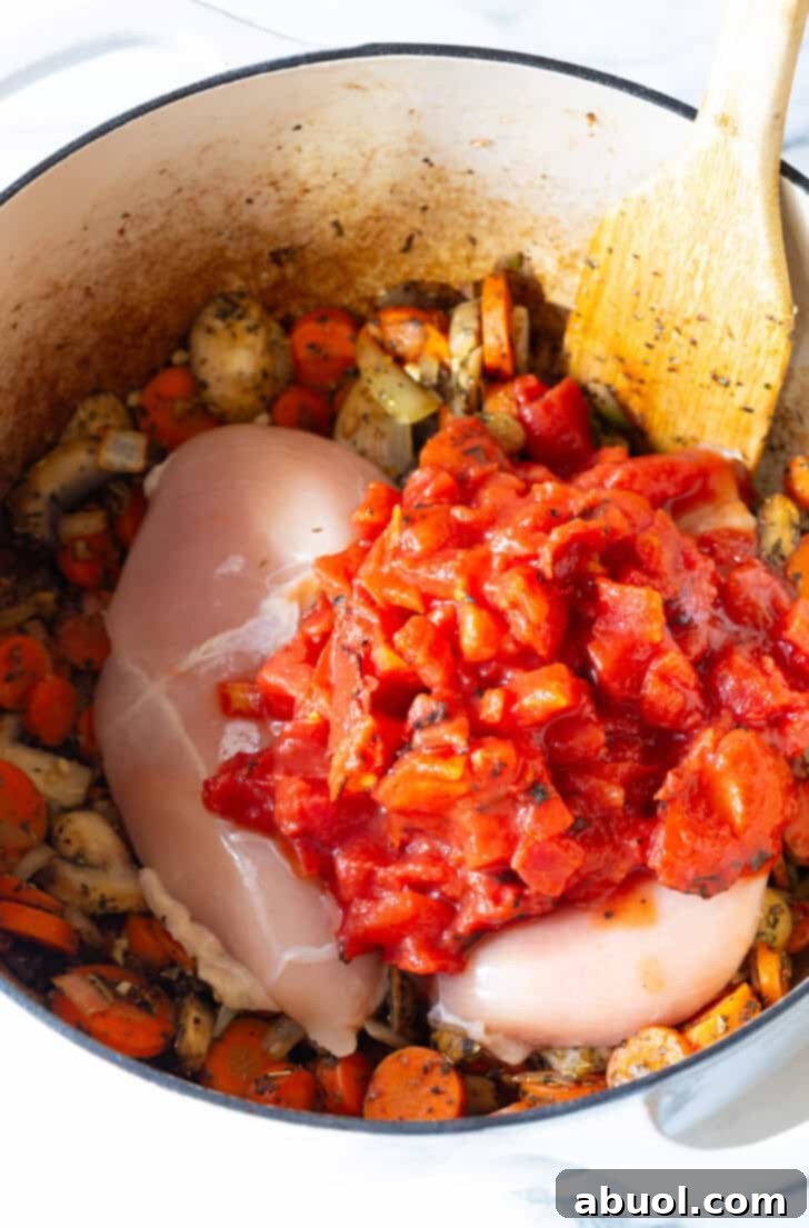 Two whole chicken breasts nestled in a large pot alongside a medley of tomatoes, zucchini, and carrots, simmering gently.