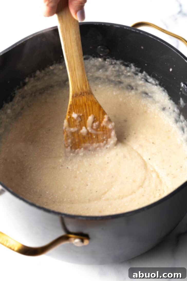 A wooden spatula stirs creamy, cheesy grits in a large black cooking pot, showcasing the smooth texture and rich consistency of the Southern staple.